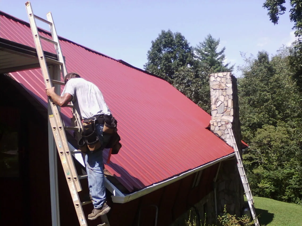 Roofer on a ladder beside a red metal roof and stone chimney — Soffit & Fascia Repair by Roofing Brothers in Allenwood